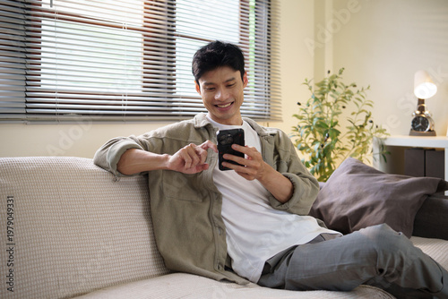 Bild auf Leinwand Young Asian man relaxing on a sofa while using a smartphone at home