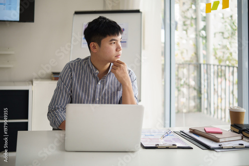 Businessman sitting at a desk with a laptop, looking out the window thoughtfully in a bright modern office