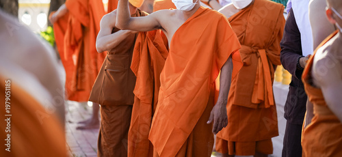 A senior monk adjusts a novice robe as hands tighten the woven belt around orange cloth in sunlight with focus on texture folds and precise fitting.