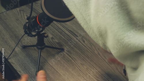 Anonymous Podcast Host In Hood Speaking Into Microphone On Wooden Desk. Close Up Overhead View Of Person Gesturing With Hands While Recording Audio Content. Professional Home Studio