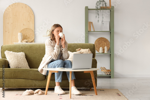 Beautiful young woman using laptop and drinking coffee on sofa in living room