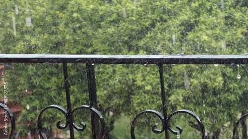 Close up of heavy rain droplets splashing on a black wrought iron balcony railing. Cinematic wet weather background with lush green trees, perfect for seasonal and moody projects.