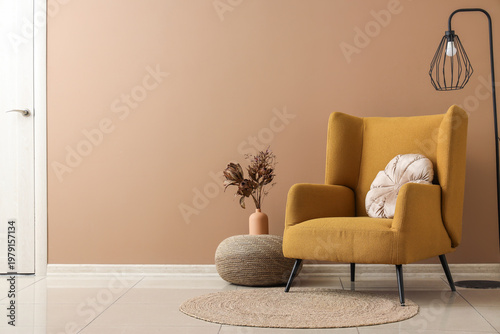 Armchair, lamp and pouf with flowers in vase near brown wall in room