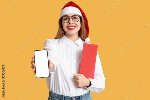 Young female real estate agent in Santa hat showing mobile phone with blank s...