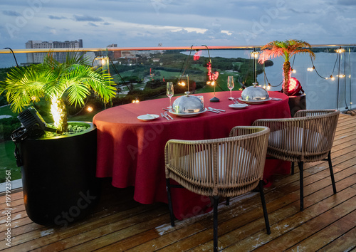 An elegant dining setup featuring a lavish table with silver cloches and ambient lighting, overlooking a picturesque landscape at dusk, creating a romantic atmosphere.