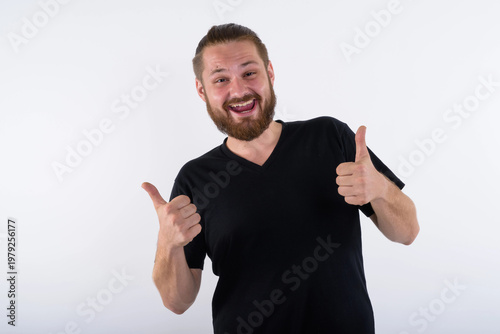 Studio portrait of a cheerful young caucasian man with a beard and hair bun wearing a black v-neck shirt. He is giving a double thumbs up gesture with a big smile on a white background.