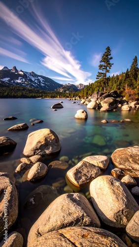 Stunning waterside landscape shows a lake with clear, turquoise water, rocky shore, and mountain backdrop under a streaky sky