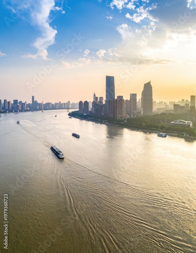 An aerial view captures a vibrant cityscape along a wide river at sunrise, showcasing modern architecture and bustling waterway traffic under a partly cloudy sky.