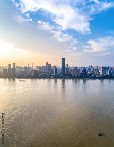 An aerial view showcases a sprawling cityscape reflected in a calm river under a bright, partly cloudy sky, highlighting the urban landscape and natural beauty.