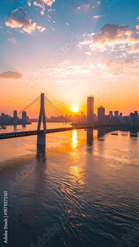 A stunning aerial view captures a modern city skyline bathed in the warm glow of a vibrant sunset, with a graceful suspension bridge spanning a tranquil river.