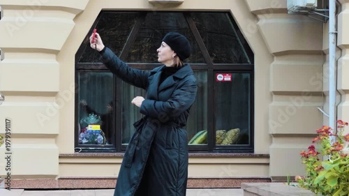 Middle-Aged Woman in Black Coat and Beret Taking Photo with Red Smartphone Outdoors