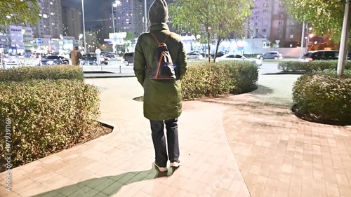 Woman in Green Winter Coat and Beanie Walking on City Sidewalk at Night with Urban Background