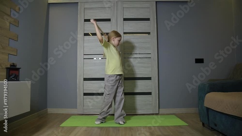 Young girl performs warm-up. Wooden stick behind shoulders. Torso rotation for flexibility. Green yoga mat, wood floor. Home interior for child fitness.