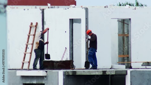 Two male construction workers performing manual labor on building site, shoveling cement or other material into mixing trough, focused on work, daytime activity