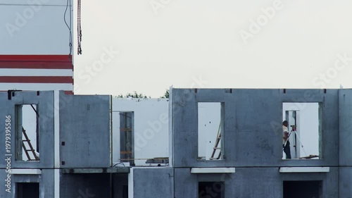 Multi story building construction shows concrete wall panels with window openings Crane lifts components for placement Surveyor operates equipment from wall opening urban development