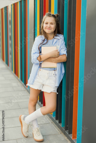 Smiling child with book leaning on colorful outdoor school wall