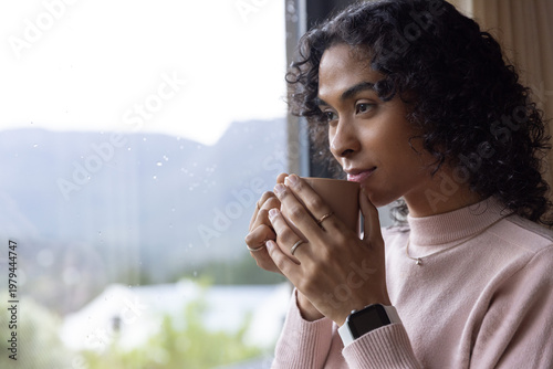 Indian nonbinary holding beige mug, wearing turtleneck and rings, gazing out wet window, copy space