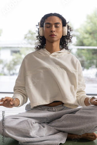 Non-binary adult meditating crosslegged on green mat wearing cream hoodie gray sweats headphones