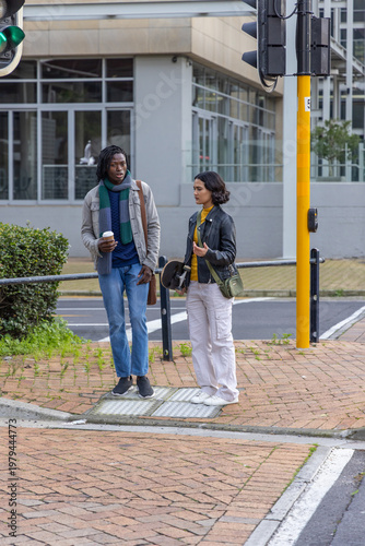 Diverse couple chatting while waiting at street crossing holding coffee cup, skateboard and scarf