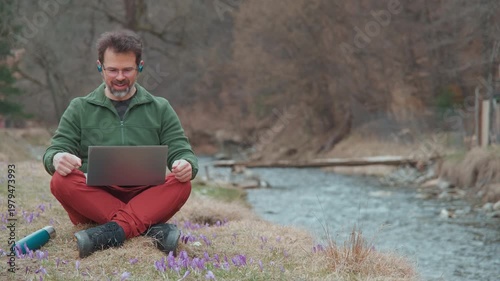 Confident senior consultant digital nomad leading a video call by a tranquil mountain river with spring flowers, cinematic static shot