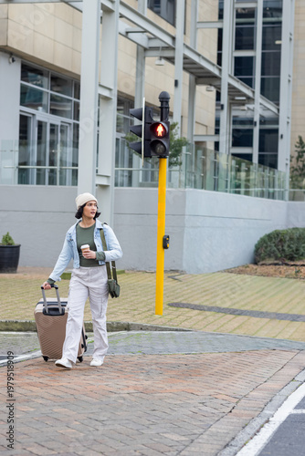 20s woman in denim jacket beanie walking across office plaza pulling rolling suitcase holding cup
