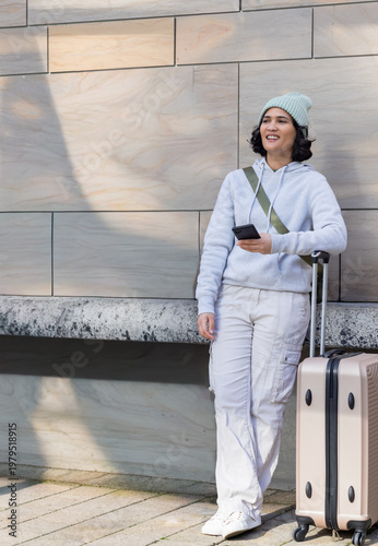 Woman standing by beige suitcase, leaning on ledge, holding phone, beanie at plaza, copy space
