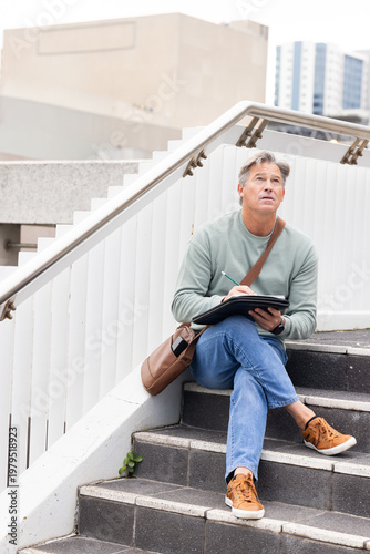 Middle-aged man sitting on outdoor concrete steps holding sketchbook and pen with satchel nearby
