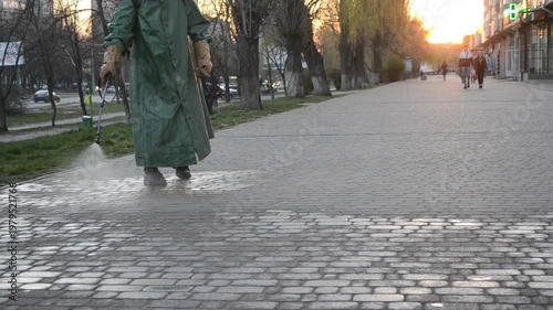 Man in airtight suit sprays disinfecting liquid chemicals antiseptic on street in city on day. Sanitation workers clean pavement, crosswalk. Sanitary measures in public place. Coronavirus pandemic