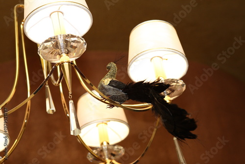 Cuadro en lienzo An elegant chandelier with gold accents and crystal droplets, featuring a decorative black peacock ornament perched on its arm