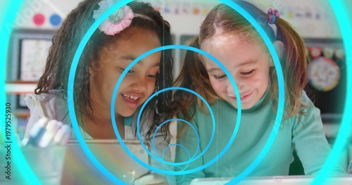 Two girls leaning over tablets tapping in classroom, generating teal rings over them for learning
