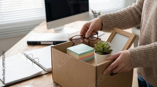 A close-up of a worker's hands placing personal items, including a small potted plant and notebooks, into a cardboard moving box on an office desk near a bright window.