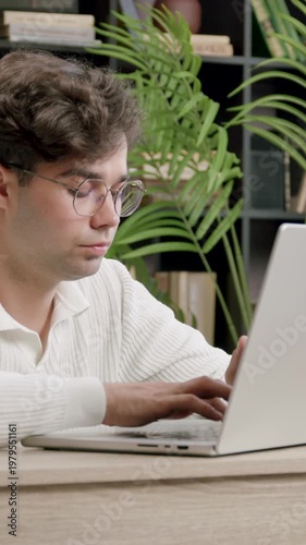 Young male student with curly hair wearing glasses types on laptop at wooden desk surrounded by books and plants in a cozy indoor study environment