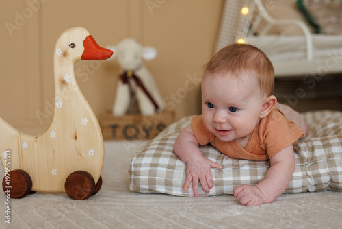 Close-up of a smiling baby lying on a checkered pillow near a wooden toy. Focus on the child's face