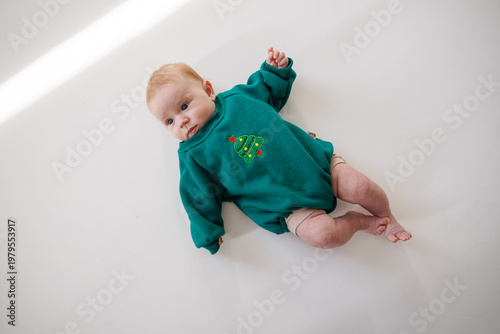 A baby lies on a white surface wearing a green sweatshirt with a Christmas tree embroidery. Top view