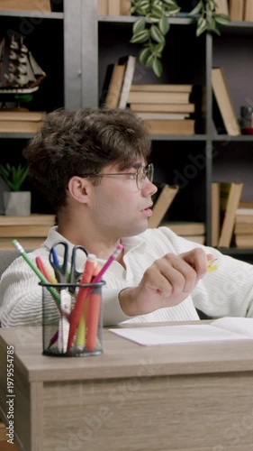 Young male student in white sweater sits at desk, holding yellow pen, contemplating notes with books and plants in the background, focused on study tasks