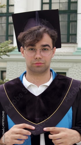 Male student in graduation gown adjusts cap while standing outdoors in front of a university building, showcasing pride and anticipation for future achievements