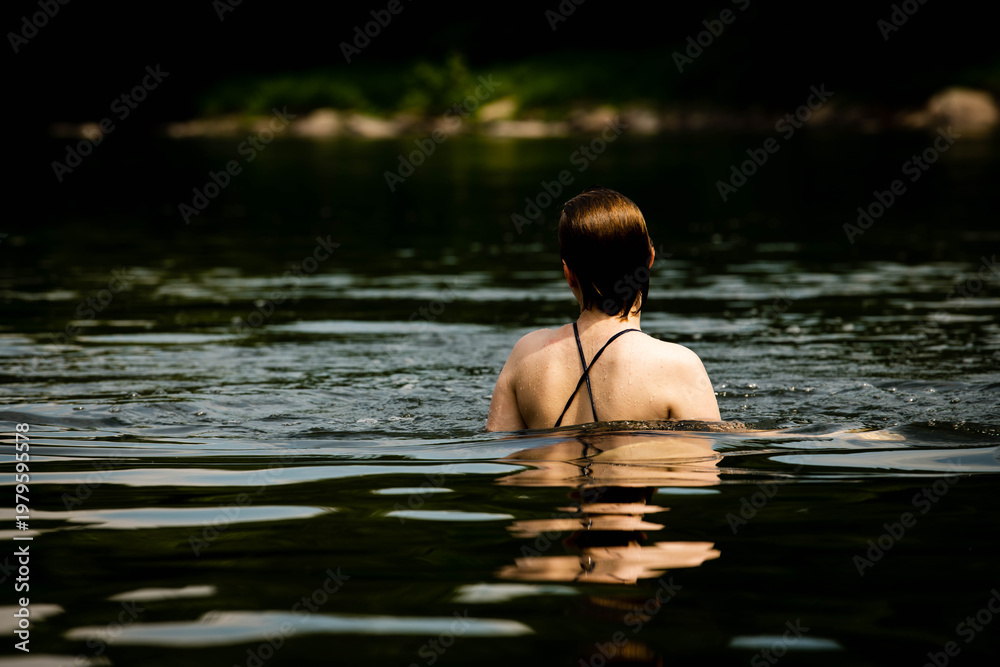 Fototapeta premium Woman from behind swimming in a dark natural lake or river, her wet hair and swimsuit strap visible above the water in a quiet and contemplative outdoor swimming scene.
