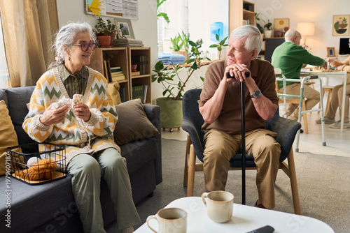Canvas Print Senior Caucasian woman knitting while senior Caucasian man resting on cane and conversing in care home lounge