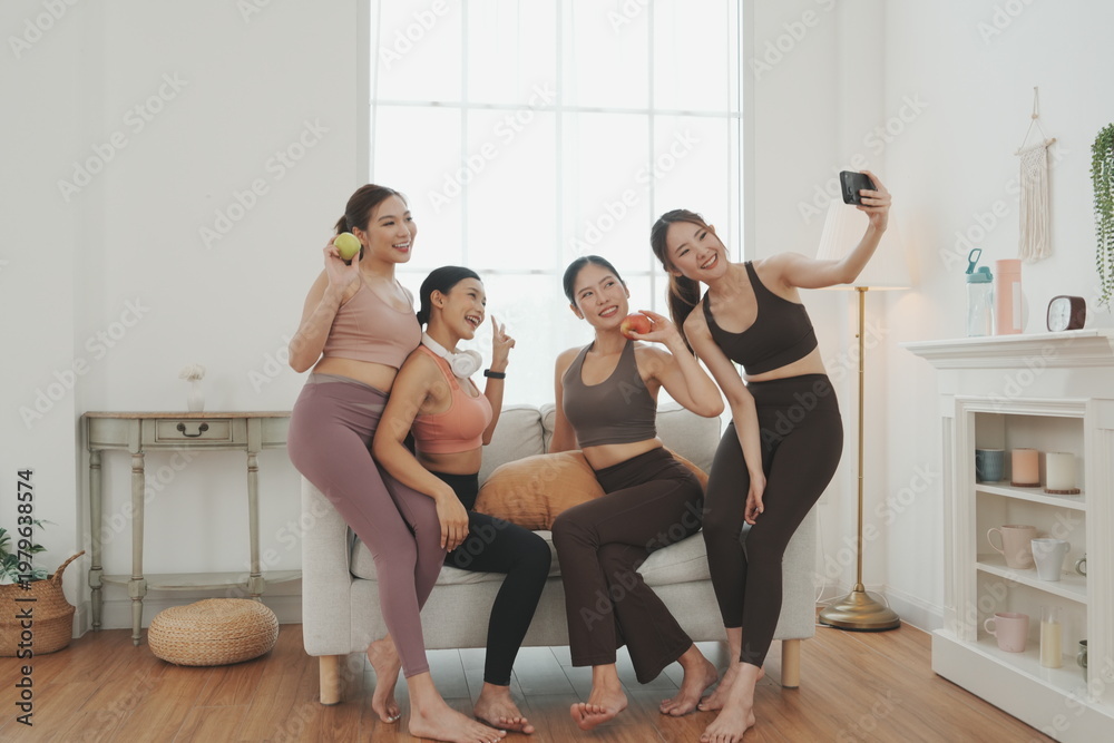 Obraz premium A group of Asian female friends, dressed in sports bras, are sitting together on a sofa after a yoga class, chatting about healthy eating, wellness concepts, and exercise. 