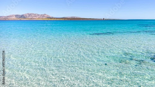 Cinematic low altitude drone flight skimming the surface of transparent turquoise water at La Pelosa beach in Sardinia showing white sand patterns and sunlight reflections on the sea