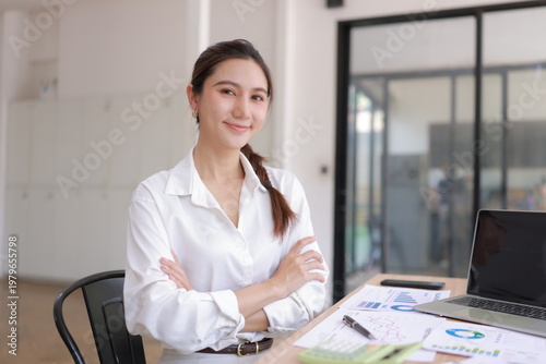Portrait of a professional Asian female accountant or entrepreneur in white shirt sitting at her workplace with laptop, financial charts, and calculator, looking at camera with a confident smile.