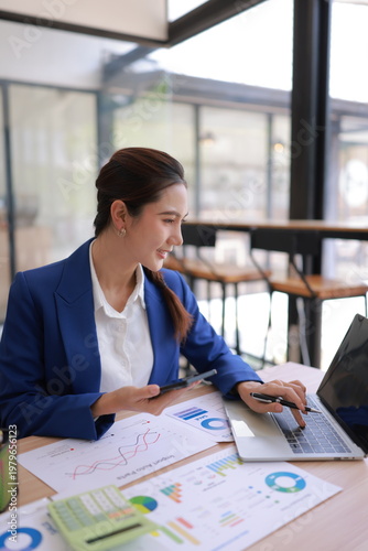 A professional Asian female executive sitting at a desk, looking at a data visualizations, graphs, and business analytics. She is holding a smartphone and has printed reports on the table.