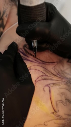 Close-up of a female tattoo artist in black latex gloves using a wireless rotary tattoo pen to ink a large, intricate floral and mandala design on a woman's upper back.