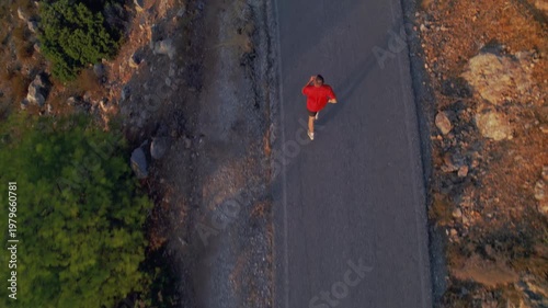 Drone tracking shot of a man running on an asphalt path near mountains. Golden hour light, soft shadows and movement create a cinematic, calm and motivational scene.