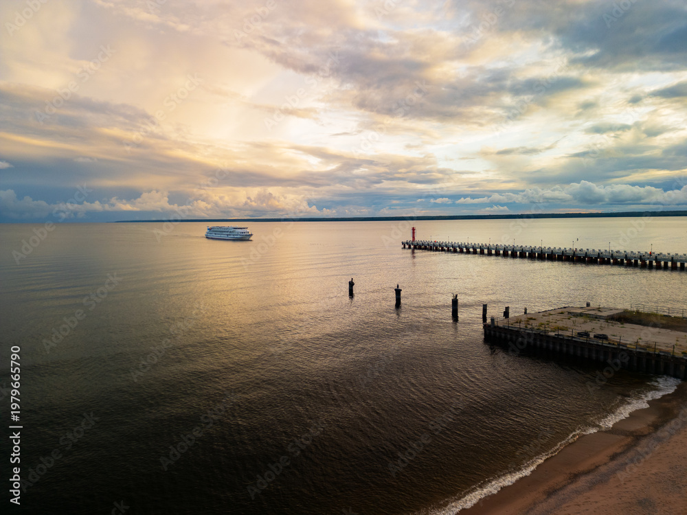 Fototapeta premium Aerial panoramic view of a cruise ship leaving the pier at sunset. Scenic northern lake coastline with cinematic sunset sky.