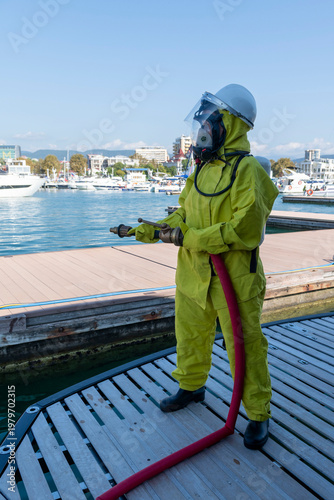 Fire drill on yacht stern: crew member in protective suit uses fire hose and breathing apparatus during training, simulating emergency firefighting on board.