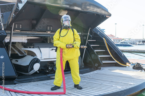 Fire drill on yacht stern: crew member in protective suit uses fire hose and breathing apparatus during training, simulating emergency firefighting on board.