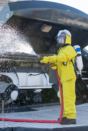 Fire drill on yacht stern: crew member in protective suit uses fire hose and breathing apparatus during training, simulating emergency firefighting on board.