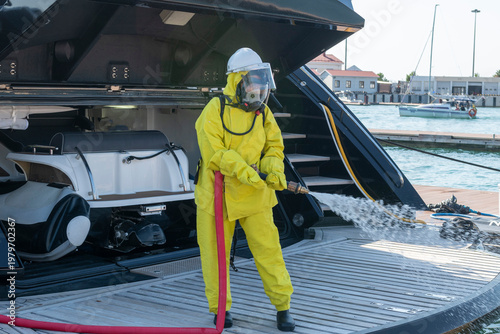 Fire drill on yacht stern: crew member in protective suit uses fire hose and breathing apparatus during training, simulating emergency firefighting on board.