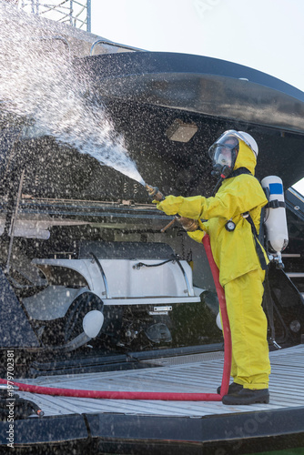 Fire drill on yacht stern: crew member in protective suit uses fire hose and breathing apparatus during training, simulating emergency firefighting on board.